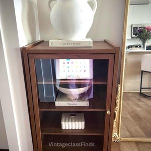 May include: A dark wood cabinet with glass doors, displaying a tablet, books, and decorative items. A white vase with handles sits atop the cabinet, with the text "The Art of Home" visible. The cabinet is next to a gold-framed mirror.