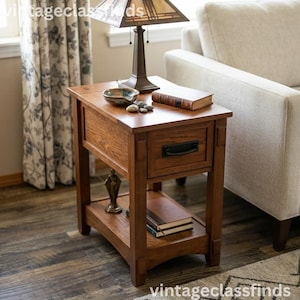 May include: A rectangular wooden end table with a medium brown finish, featuring a single drawer with a black metal handle and an open lower shelf. The table surface holds a lamp, a small bowl, and a book, while the lower shelf holds two books.