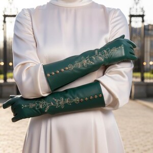 Könnte beinhalten: Smaragdgrüne Lederhandschuhe mit floraler Stickerei und goldfarbenen Knöpfen. Die Handschuhe reichen bis zum Ellbogen und werden über einem weißen Kleid getragen. Die Handschuhe haben ein klassisches Design.