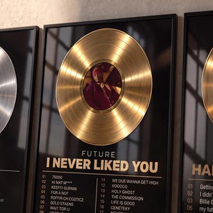 Könnte beinhalten: Drei gerahmte Wandbilder mit metallischen Schallplatten. Der linke Druck zeigt eine silberne Platte für Drakes Views. Der mittlere Druck zeigt eine goldene Platte für Futures I Never Liked You. Der rechte Druck zeigt eine goldene Platte für Billie Eilishs Happier Than Ever, jeweils mit Titellisten.