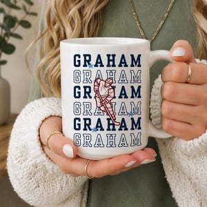 Może przedstawiać: Biały ceramiczny kubek z napisem "GRAHAM" w granatowych literach blokowych i grafiką hokeisty w kolorze czerwonym i białym. Kubek ma biały uchwyt i jest ozdobiony niebieskimi gwiazdami.