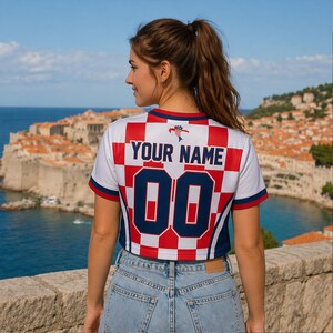 Op de afbeelding: Een wit, rood en blauw cropped shirt met de tekst "YOUR NAME" en het nummer "00" op de rug. Het shirt heeft een geruit patroon en wordt gedragen met een blauwe jeans. De achtergrond toont een kuststad en blauw water.