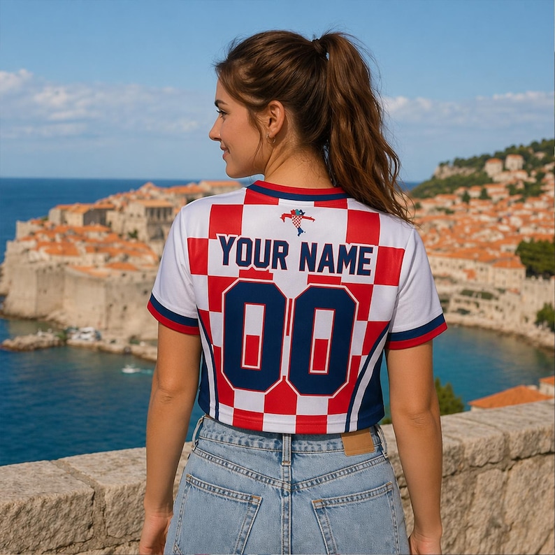 Op de afbeelding: Een wit, rood en blauw cropped shirt met de tekst "YOUR NAME" en het nummer "00" op de rug. Het shirt heeft een geruit patroon en wordt gedragen met een blauwe jeans. De achtergrond toont een kuststad en blauw water.