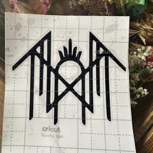 May include: Black vinyl decal featuring a stylized design with a central hand-like shape, an X, and vertical lines. The decal is on a grid-patterned sheet with the text "cricut Transfer Tape".