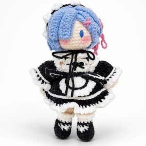 May include: A handmade crochet doll of a character with blue hair, wearing a black and white maid outfit. The doll has a light skin tone, pink accents, and black shoes. The doll is standing against a white background.