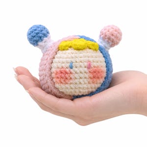 May include: A handmade, crocheted plush toy in the shape of a round character. The toy features a cream-colored face with pink cheeks, blue and pink accents, and yellow details. The toy is held in a hand.