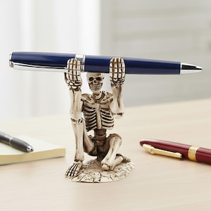 May include: A novelty pen holder in the shape of a skeleton kneeling and holding a blue pen. The pen holder is off-white with a detailed skeleton design. A dark blue pen with silver accents is held by the skeleton. Additional pens are visible in the background.