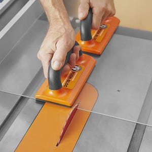 May include: Two orange push blocks with black handles are used to guide a piece of clear material through a table saw. The saw blade is visible, and the table is grey.