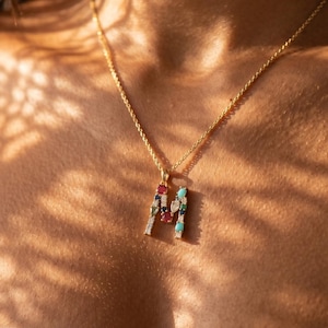 Pode incluir: Um colar de corrente de corda dourada com um pingente em forma de letra M. O pingente é adornado com uma variedade de pequenas pedras preciosas multicoloridas, incluindo pedras vermelhas, azuis e transparentes cravadas no metal.