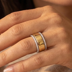 Puede incluir: Un anillo ancho de oro con una banda de pequeñas piedras transparentes a cada lado. El anillo está en un dedo, con un fondo suave.