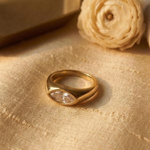 Könnte beinhalten: Ein goldener Ring mit einem klaren, marquiseförmigen Edelstein. Der Ring liegt auf einem cremefarbenen Stoff, mit einem goldfarbenen Tablett und einer cremefarbenen Blume im Hintergrund. Ein auffälliger Ring.