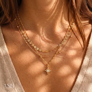 Könnte beinhalten: Ein mehrschichtiges goldenes Halsketten-Set. Die oberste Schicht hat kleine, runde, funkelnde Verzierungen. Die mittlere Schicht hat eine zarte Kette mit kleinen, flachen, kreisförmigen Anhängern. Die untere Schicht hat einen Sternanhänger.