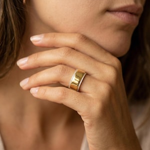 May include: A broad gold ring featuring a row of small, clear gemstones. The ring has a polished, reflective surface and is worn on a finger. The ring is a piece of jewellery.