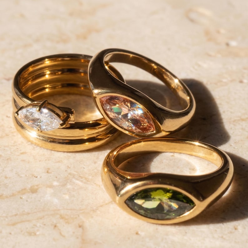 May include: Three gold rings with oval-shaped gemstones. One ring features a clear gemstone, another a peach-colored stone, and the third a green stone. The rings are displayed on a light-colored surface.