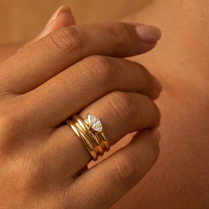 May include: A set of gold rings, including a band with a pear-shaped diamond. The rings are stacked on a finger, showcasing a minimalist and elegant jewelry design. The gold has a polished finish, reflecting light.
