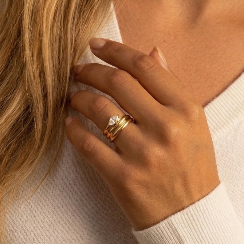 May include: A close-up of a hand wearing a set of gold rings. The rings include a band, a stack of two plain bands, and a ring with a clear, triangular-cut gemstone. The hand is resting on a white sweater.