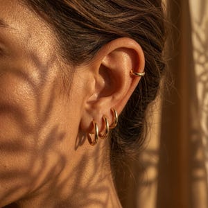 Peut inclure: Gros plan sur une oreille ornée de boucles d'oreilles créoles dorées et d'un ear cuff doré. Trois créoles dorées de tailles différentes sont visibles. L'ear cuff est en haut de l'oreille. L'éclairage crée des ombres.