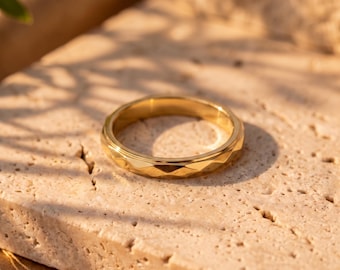 Puede incluir: Un anillo de metal en tono dorado que presenta un patrón de superficie geométrico y facetado alrededor de la banda exterior. El anillo tiene un acabado pulido y reflectante, con una forma circular y una serie de cortes angulares repetidos.