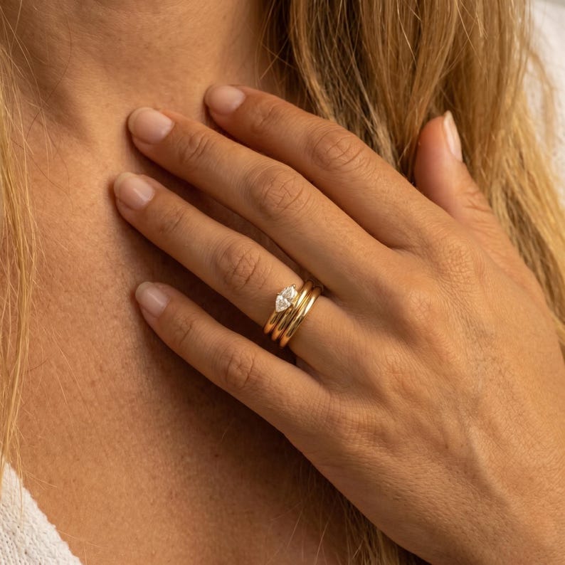 May include: A set of three gold rings. The top ring features a pear-shaped diamond. The other two rings are simple gold bands. The rings are stacked on a finger.