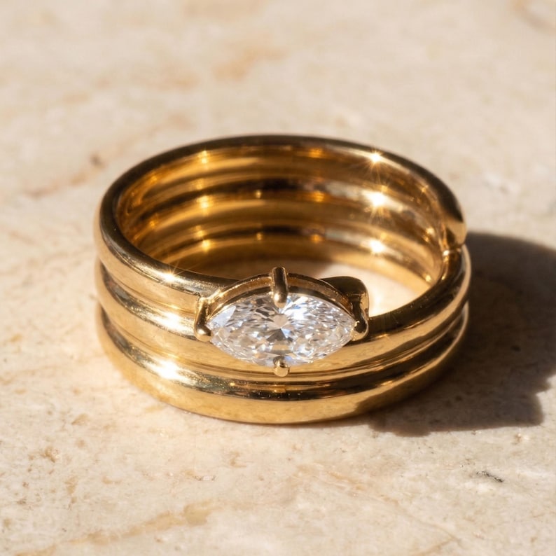 May include: A gold ring with a marquise-cut diamond. The ring has a unique design with multiple bands and a central diamond setting. The ring is displayed on a light-colored surface, showcasing its polished finish and intricate details.
