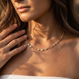 Peut inclure: Un collier en or au design délicat avec une série de petites pierres précieuses scintillantes et des formes géométriques. Le collier est porté autour du cou, et une bague en or est visible sur le doigt. L'esthétique générale est élégante et raffinée.