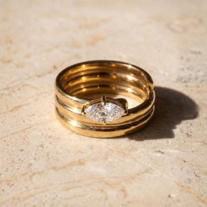 May include: A gold ring with a marquise-cut diamond set in the center. The ring has a multi-layered band design. The ring is displayed on a light-colored, textured surface.
