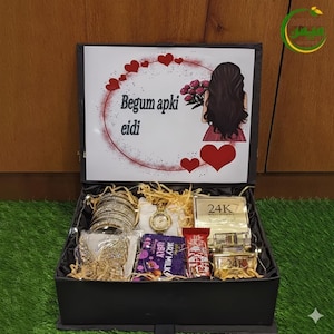 Può includere: Una scatola regalo nera piena di vari oggetti, tra cui braccialetti d'oro, un orologio, barrette di cioccolato e piccole scatole color oro. Il coperchio della scatola presenta un disegno di una donna e il testo "Begum apki eidi" con accenti a cuore.
