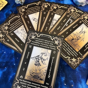 May include: A deck of tarot cards with black and gold designs, featuring illustrations and text. The central card, "The Fool," displays a figure with the text "Spread your wings and fly." Other cards show astrological symbols and keywords.