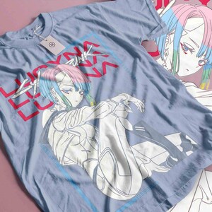 May include: A pale blue t-shirt with an anime-style character graphic with pink and blue hair. The shirt features the text "LUCKY" in red and "V" in a blue box. A tag is attached to the shirt.