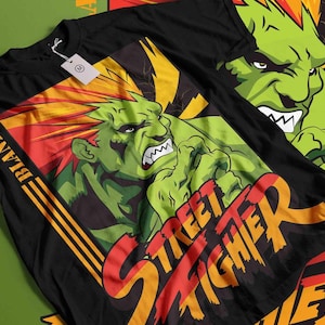 Puede incluir: Camiseta negra con un personaje verde con pelo rojo y amarillo y las palabras "Street Fighter" en rojo y naranja. El personaje está en una pose de lucha con una expresión feroz. La camiseta tiene una etiqueta.