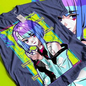 May include: A navy blue t-shirt featuring a vibrant anime-style illustration of a female character with blue and purple hair. The design includes the word "LUCY" and other text in a neon green and blue color scheme. The shirt has a tag attached.