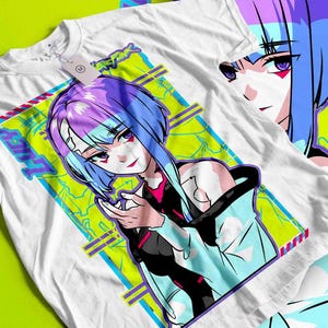 May include: White t-shirt featuring a colorful anime-style illustration of a female character with blue and purple hair. The design includes the word "LUCY" and other text within a geometric frame. A second shirt with a partial image is visible.