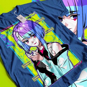 May include: A blue t-shirt featuring a vibrant anime-style illustration. The graphic showcases a female character with blue and purple hair, set against a neon green and yellow background. The word "LUCY" is visible on the design, along with the text "CYBERPUNK".