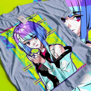 May include: A light blue t-shirt featuring a vibrant anime-style illustration of a female character with blue and purple hair. The design includes the text "LUCY" and "CYBERPUNK" in a neon green and blue color scheme.