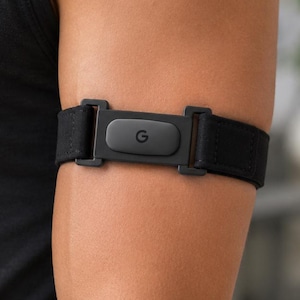 May include: A black heart rate monitor sensor with a central G logo, attached to an adjustable elastic strap. The device features a rectangular plastic housing with integrated clips that secure the fabric band for placement on the upper arm.