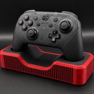 May include: A black and gray Nintendo Switch Pro Controller with a camouflage pattern, resting on a red and black charging dock. The controller has multiple buttons and joysticks. The dock has a ribbed design.