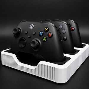 May include: A white charging station for Xbox controllers, holding three black gamepads. The controllers have coloured buttons and joysticks. The station has a ridged design and a black interior.