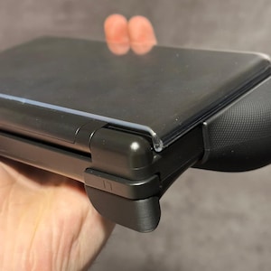 May include: A dark grey handheld gaming console with a clear screen protector. The console features a textured grip and an "L1" button. The device is held in a hand.