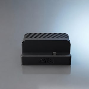 May include: A matte black rectangular charging dock featuring a central USB-C connector, a grooved top surface for ventilation, and the embossed AYN logo on the front base. The device is designed for electronic hardware support and connectivity.