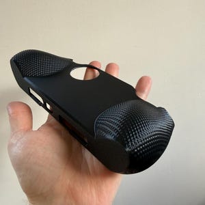 Puede incluir: Una funda negra para consola de juegos portátil con agarres texturizados. La funda tiene una abertura circular y varios recortes laterales. Los agarres tienen un patrón repetitivo de pequeños puntos en relieve.