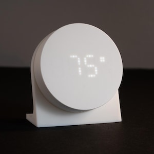 May include: A white, circular smart thermostat displaying 75°F (23.8°C). The device has a digital display and is mounted on a white, rectangular base. It is designed for home climate control and temperature regulation.