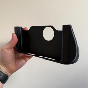 May include: A black camera grip accessory featuring a contoured handle with a textured surface on the right side, a central circular cutout, and a rectangular frame designed to attach to the base of a camera body for improved handling.