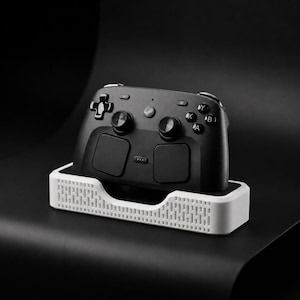 May include: A black wireless game controller with a white charging dock. The controller has two analog sticks, a directional pad, and four action buttons. The dock is rectangular with a textured pattern.