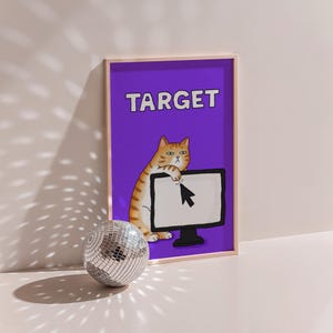 May include: A framed print featuring a cartoon cat with orange and white stripes, leaning on a computer monitor with a cursor. The word "TARGET" is displayed in white letters on a purple background. A disco ball sits in front of the print.