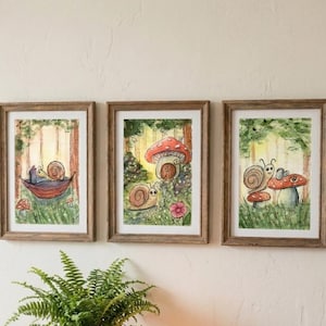 May include: Three framed watercolor art prints featuring whimsical snail illustrations in a forest setting. Each print has a wooden frame. The snails are depicted with mushrooms, hammocks, and flowers, in a vibrant color palette of greens, reds, and browns.