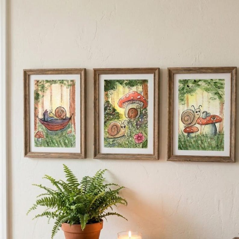 May include: Three framed watercolor art prints featuring whimsical snail illustrations in a forest setting. Each print has a wooden frame. The snails are depicted with mushrooms, hammocks, and flowers, in a vibrant color palette of greens, reds, and browns.