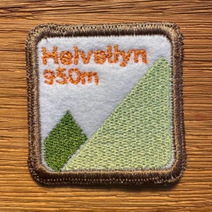 May include: A square embroidered patch with a white background, a dark brown stitched border, and orange text reading Helvellyn 950m. The design features a large light green triangular mountain shape and a smaller dark green triangular tree shape.