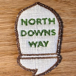 May include: A white embroidered patch with a brown stitched border, shaped like a signpost. The text NORTH DOWNS WAY is stitched in green thread across the centre of the patch. The design features a pointed top and a rectangular base section.