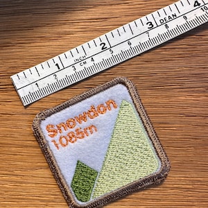May include: Embroidered square patch with the text "Snowdon 1085m" in orange, and a mountain graphic in green and light green. A ruler with both inches and centimeters is visible.