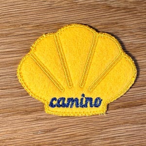 May include: A yellow felt patch shaped like a scallop shell with embroidered edges and radial lines. The word "camino" is stitched in dark blue thread across the bottom centre of the shell. The patch has a textured surface and a finished border.